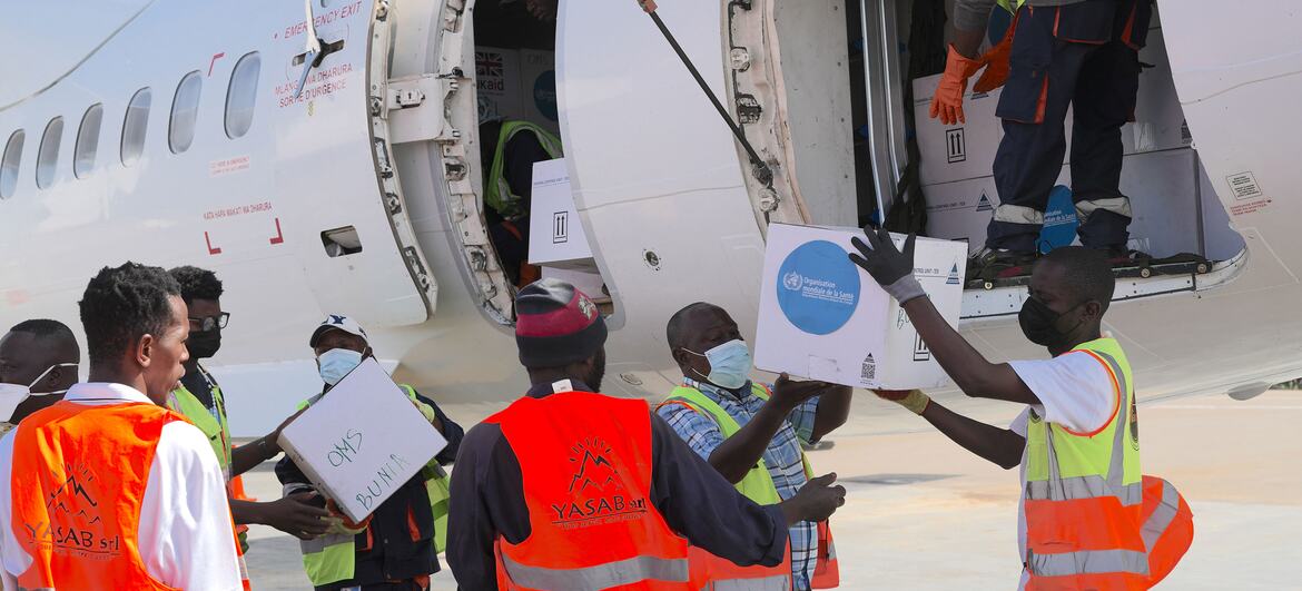 Men in safety vests and masks handle boxes with the WHO logo, loading them onto an aircraft at an airport.