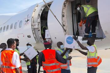 Men in safety vests and masks handle boxes with the WHO logo, loading them onto an aircraft at an airport.