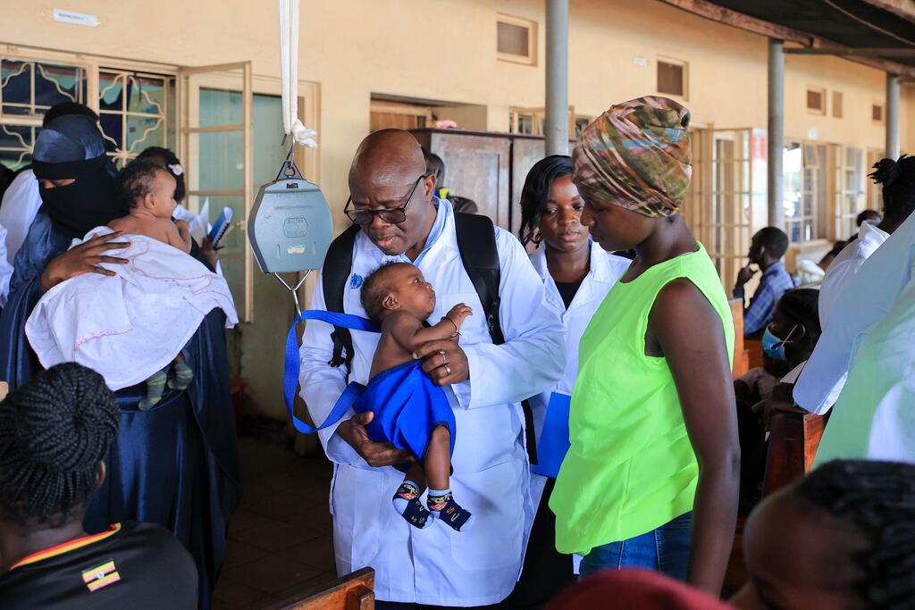 Mtaalamu wa afya mwenye kanzu nyeupe anapima uzito wa mtoto mchanga kwenye kigawanyo cha hospitali, akiwa amezungukwa na wafanyakazi wengine wa afya na wagonjwa.