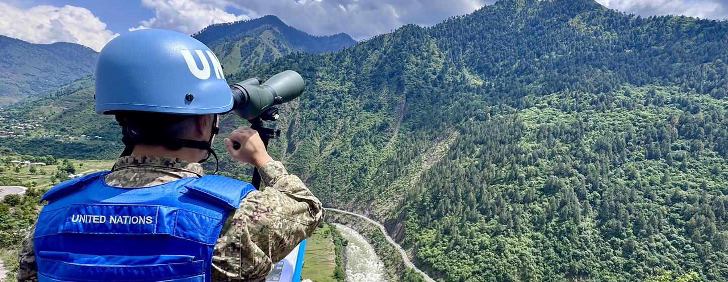 Un guardián de la paz de la ONU con casco y chaleco azules observa un paisaje montañoso a través de binoculares.