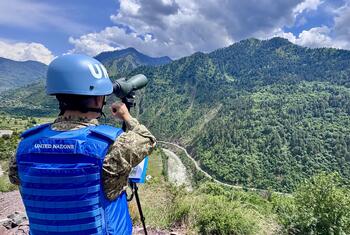 Un guardián de la paz de la ONU con casco y chaleco azules observa un paisaje montañoso a través de binoculares.