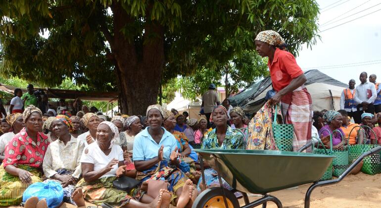 Um grupo de mulheres em Moçambique senta-se no chão debaixo de uma árvore enquanto uma mulher de camisa vermelha e saia estampada fica com uma carrinha verde, distribuindo cestos verdes de ajuda.