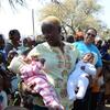 Una mujer en Mozambique sostiene a dos bebés, uno con un traje de rayas rosas y otro de color blanco, mientras que una mujer con una camisa azul de UNICEF se encuentra a su lado, rodeada de otros niños y adultos.