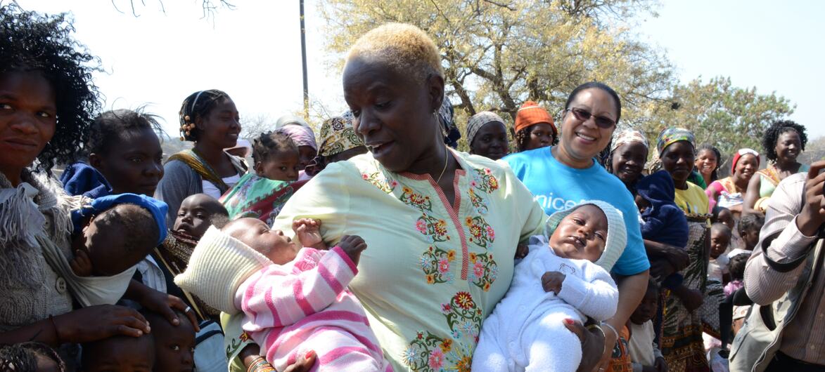 Una mujer en Mozambique sostiene a dos bebés, uno con un traje de rayas rosas y otro de color blanco, mientras que una mujer con una camisa azul de UNICEF se encuentra a su lado, rodeada de otros niños y adultos.