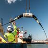 Um trabalhador com um colete de alta visibilidade e chapéu direciona a implantação de um grande cabo submarino de um navio em um porto de Porto, Portugal, durante a Cúpula Internacional de Resiliência de Cabos Submarinos de 2026.