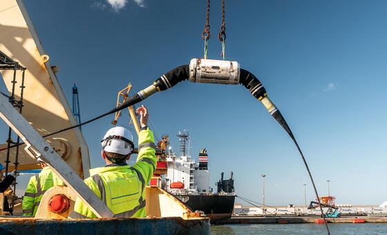 Um trabalhador com um colete de alta visibilidade e chapéu direciona a implantação de um grande cabo submarino de um navio em um porto de Porto, Portugal, durante a Cúpula Internacional de Resiliência de Cabos Submarinos de 2026.