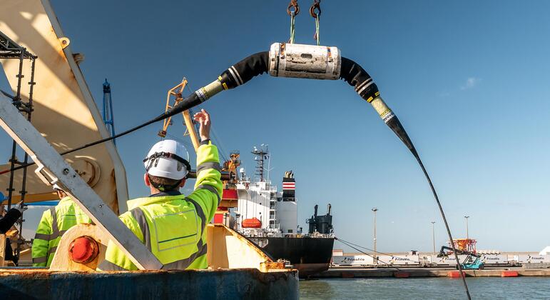 Работник в жилете с высокой видимостью и твердой шляпой направляет развертывание большого подводного кабеля с корабля в порту Порту, Португалия, во время Международного саммита устойчивости подводных кабелей 2026 года.