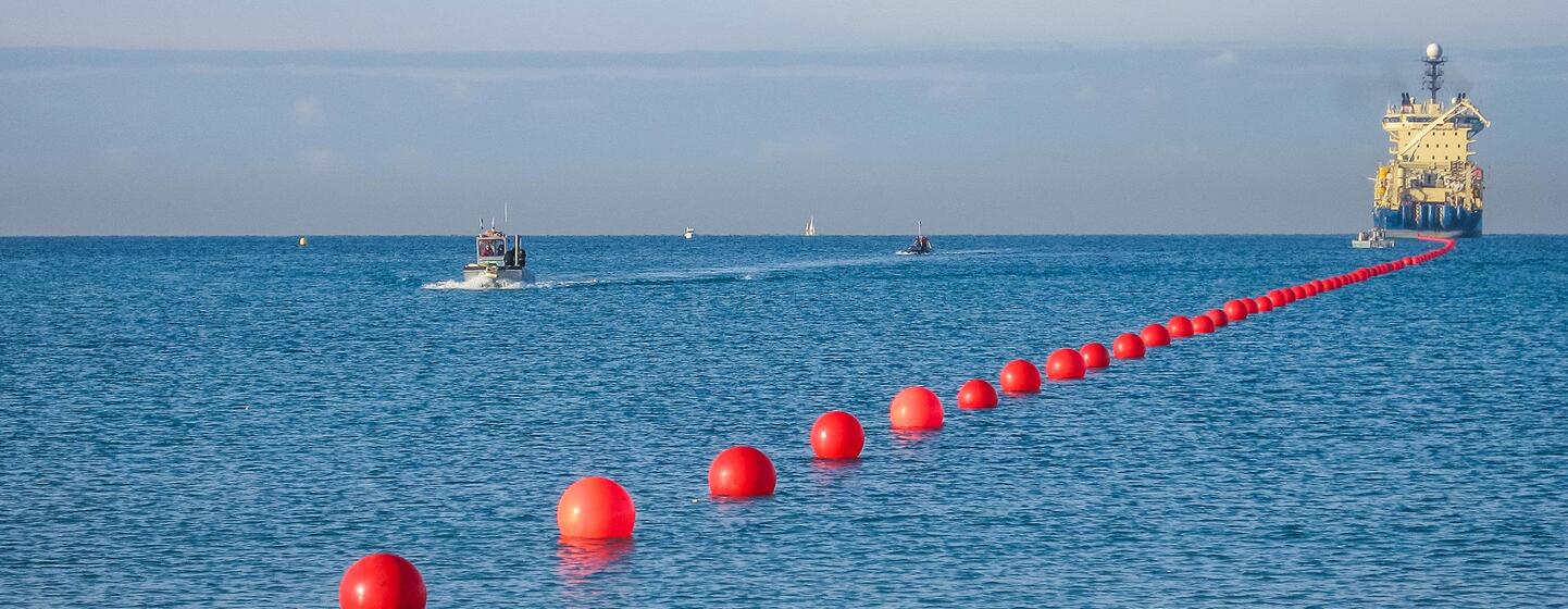 Una línea de marcadores de boyas rojas en el océano, con un buque de colocación de cables y pequeñas embarcaciones en la distancia, representando la Cumbre Internacional de Resiliencia de Cables Submarinos 2026.