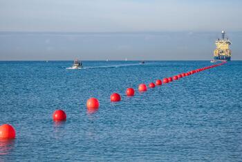 Una línea de marcadores de boyas rojas en el océano, con un buque de colocación de cables y pequeñas embarcaciones en la distancia, representando la Cumbre Internacional de Resiliencia de Cables Submarinos 2026.