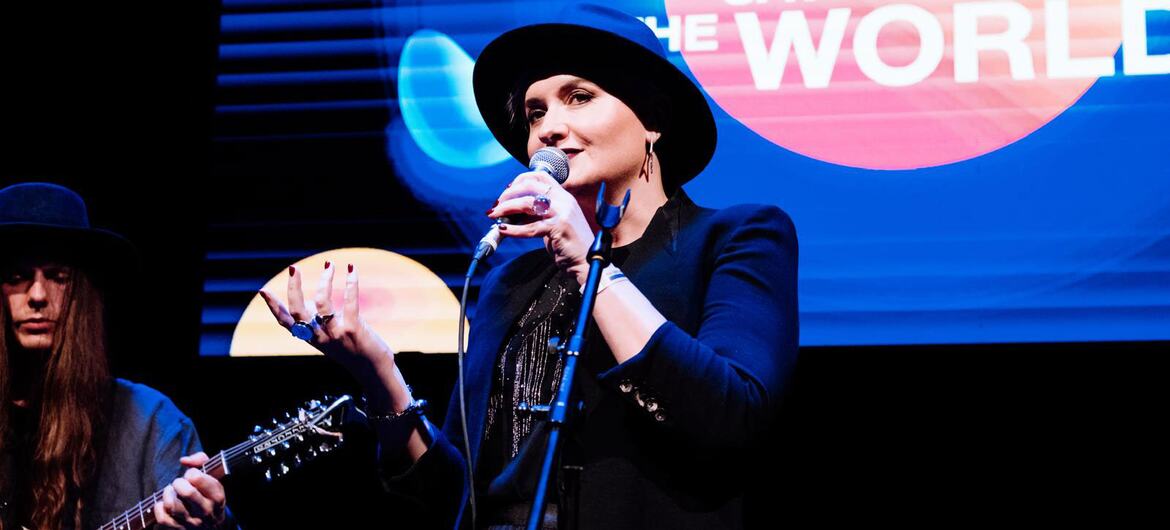 Maria Semushkina se produit sur scène au festival « Music Saves the World », chantant dans un microphone tandis qu'un guitariste joue à côté d'elle.