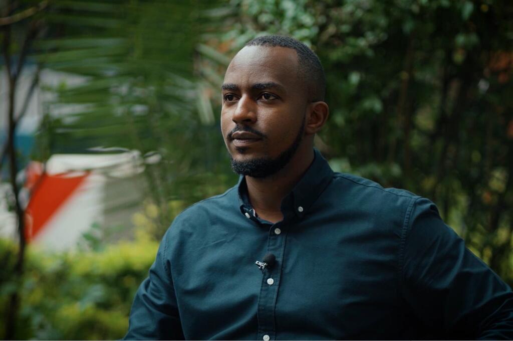 Mwanamume wa Rwanda mwenye ndevu amevaa kanzu nyeusi, akizungumza wakati wa mahojiano nje ya kijani kibichi nyuma.