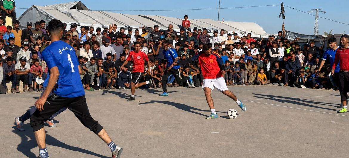 En un campo de refugiados se juega un partido de fútbol en un terreno polvoriento, con una gran multitud de espectadores observando desde las gradas. Se aprecian tiendas de campaña al fondo.