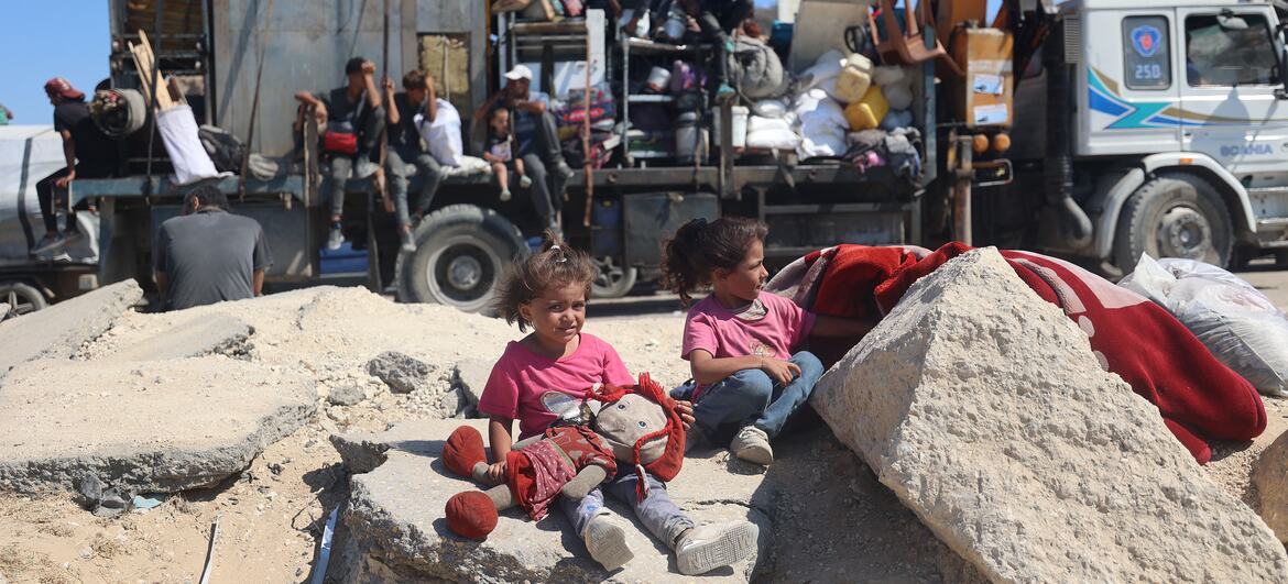 Niños palestinos sentados sobre los escombros en primer plano, con un gran camión que transporta a familias desplazadas en el fondo durante una evacuación en Gaza.