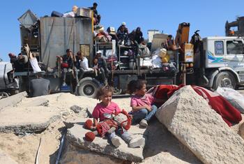 Palestinian children sit on rubble in the foreground, with a large truck carrying displaced families in the background during an evacuation in Gaza.