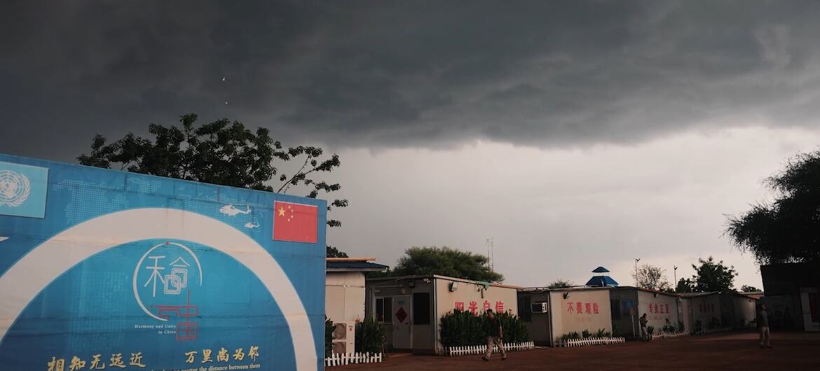 在黑暗且暴风雨的天空下，中国联合国维和基地的临时住房单元。前景中，一面巨大的蓝色墙壁上印有联合国标志、中国国旗以及汉字。