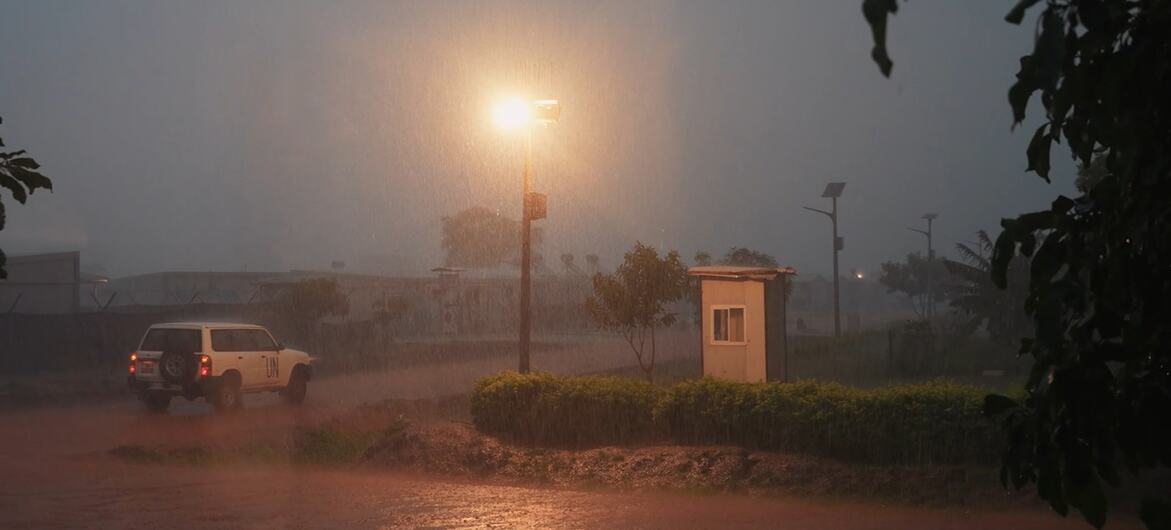 一辆白色 SUV 在暴雨期间行驶于一条被淹的道路上，旁边是一座小型哨所，场景由一盏路灯照亮。