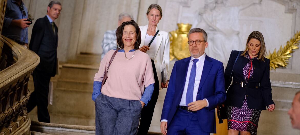 Um grupo de pessoas formalmente vestidas, incluindo uma mulher com um top rosa e um homem com um terno azul, descendo uma grande escada dentro de um edifício.