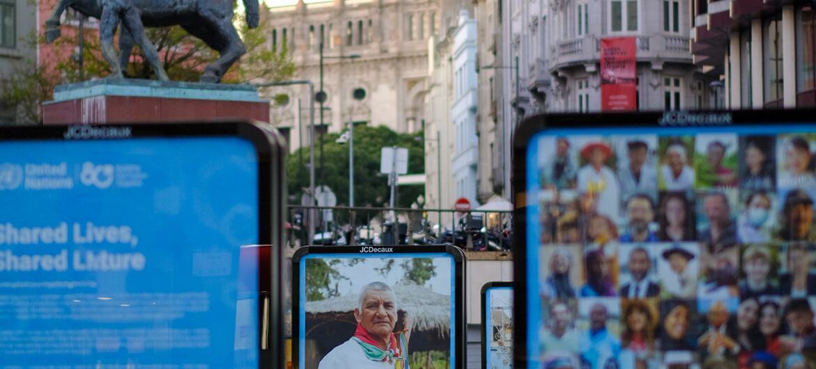 Uma cena de rua em Lisboa, Portugal, com exibições digitais para a celebração da ONU80. Uma tela mostra o texto 'Vidas compartilhadas, futuro compartilhado' com o logotipo da ONU. Outra tela mostra um retrato de um homem indígena idoso. Uma terceira tela mostra uma colagem de diversos rostos. No fundo, uma estátua de bronze de um cavalo e cavaleiro está em um sofá, com edifícios históricos alinhados na rua.