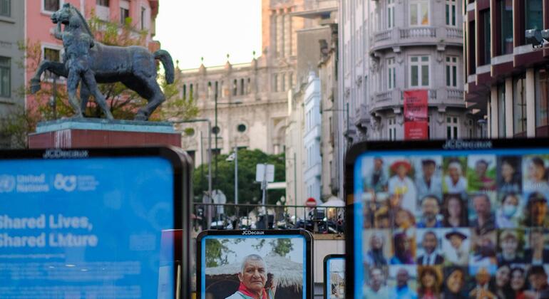 Uma cena de rua em Lisboa, Portugal, com exibições digitais para a celebração da ONU80. Uma tela mostra o texto 'Vidas compartilhadas, futuro compartilhado' com o logotipo da ONU. Outra tela mostra um retrato de um homem indígena idoso. Uma terceira tela mostra uma colagem de diversos rostos. No fundo, uma estátua de bronze de um cavalo e cavaleiro está em um sofá, com edifícios históricos alinhados na rua.