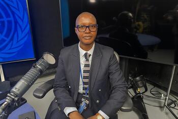Picha ya mwanasheria wa kike wa Kenya akiwa ameketi katika studio ya kitaalamu akiwa na kipaza sauti na skrini ya nembo ya Umoja wa Mataifa nyuma.