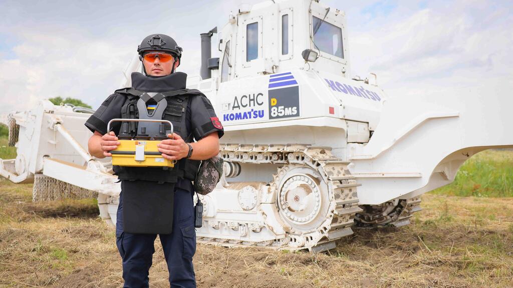 A Ukrainian deminer in full protective gear and a helmet stands in a field holding a detection device, with a large white armored Komatsu bulldozer in the background. The scene depicts landmine clearance operations in Ukraine, supported by the UNDP.