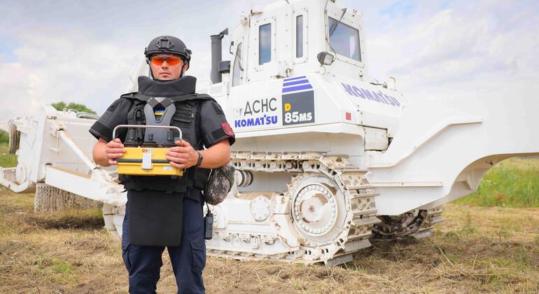A Ukrainian deminer in full protective gear and a helmet stands in a field holding a detection device, with a large white armored Komatsu bulldozer in the background. The scene depicts landmine clearance operations in Ukraine, supported by the UNDP.