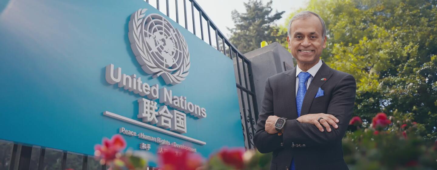 一名穿着黑色西装和蓝色领带的男子, 微笑地站在中国的蓝色联合国标志前, 标志着联合国的标志和英语和中文文字.