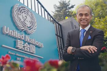 一名穿着黑色西装和蓝色领带的男子, 微笑地站在中国的蓝色联合国标志前, 标志着联合国的标志和英语和中文文字.