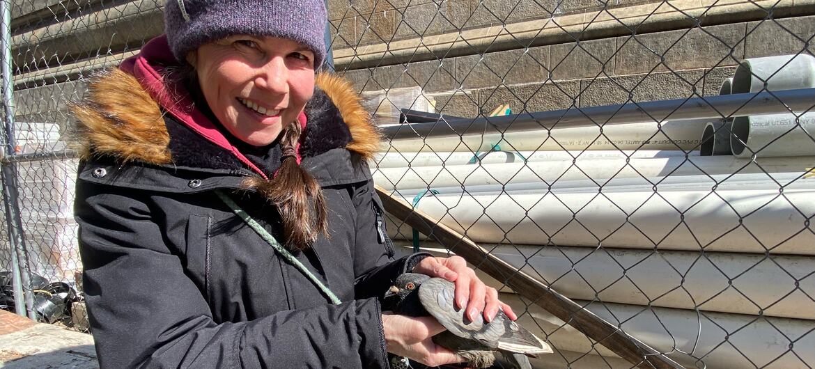 Catherine Quayle smiles while holding a pigeon in her hands, sitting outdoors near a chain link fence and stacked pipes.