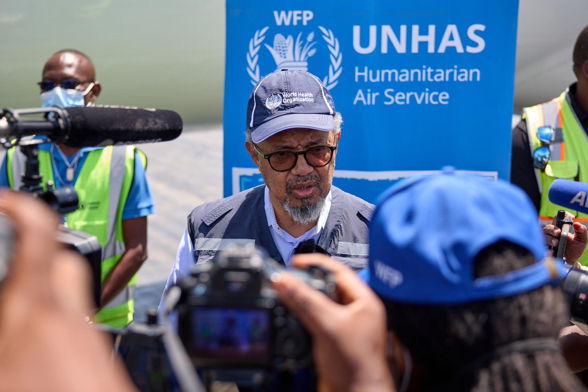 WHO Director-General Tedros Adhanom Ghebreyesus speaks at a press event, with a WFP UNHAS banner in the background and media personnel filming.