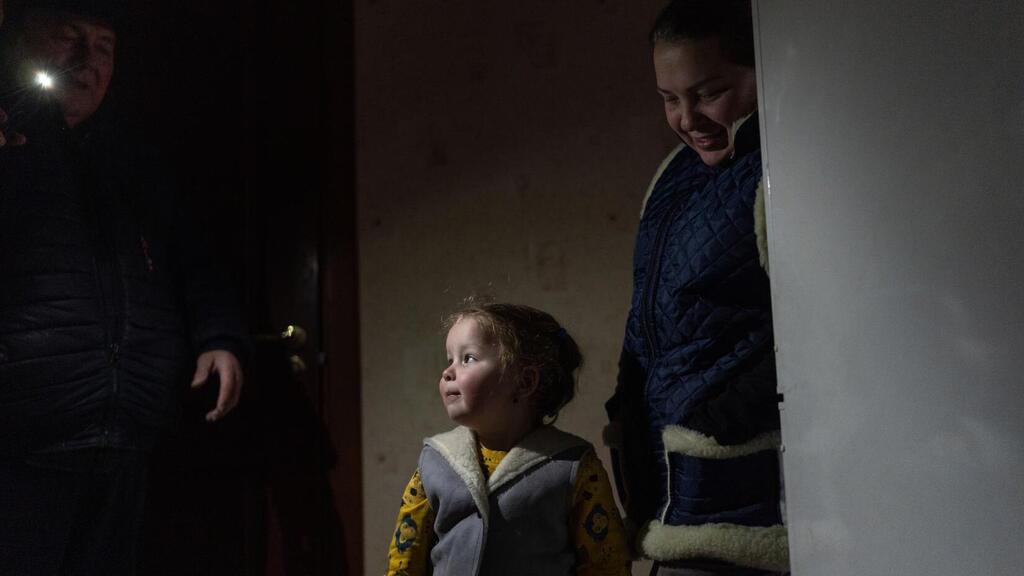 Un niño pequeño en ropa cálida de invierno se encuentra en un cuarto poco iluminado en Ucrania durante el invierno entre dos adultos, uno con una linterna en la mano.