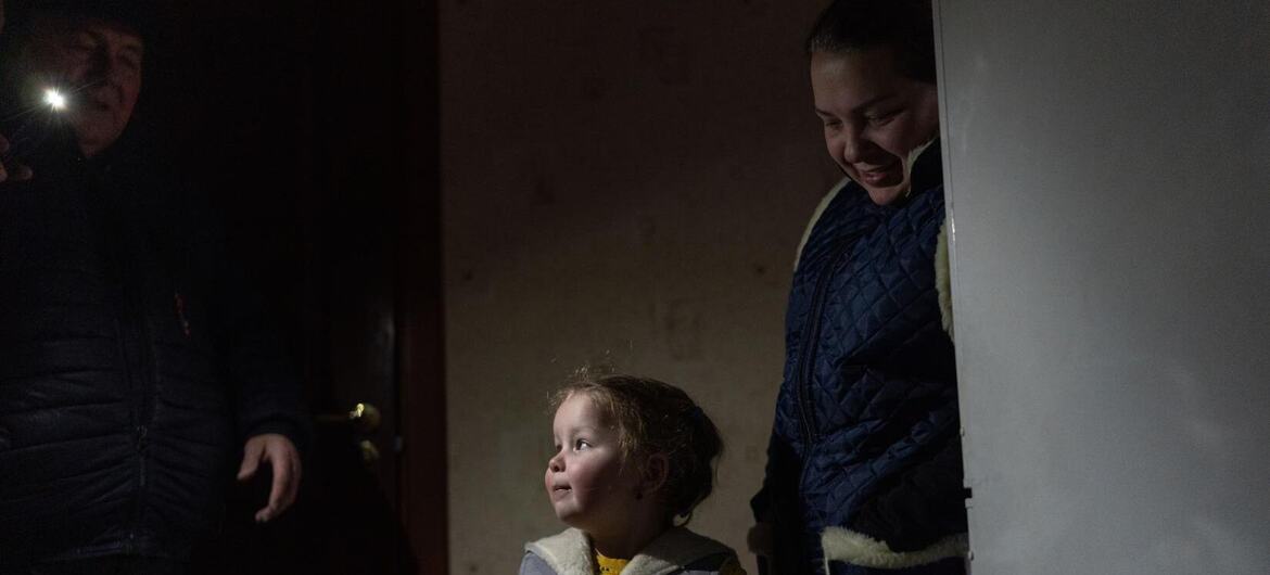 Маленький ребенок в теплой зимней одежде стоит в помещении между двумя взрослыми, один из которых держит фонарик, в слабо освещенной комнате в Украине зимой.