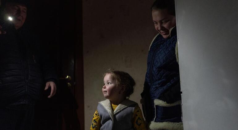 Un niño pequeño en ropa cálida de invierno se encuentra en un cuarto poco iluminado en Ucrania durante el invierno entre dos adultos, uno con una linterna en la mano.