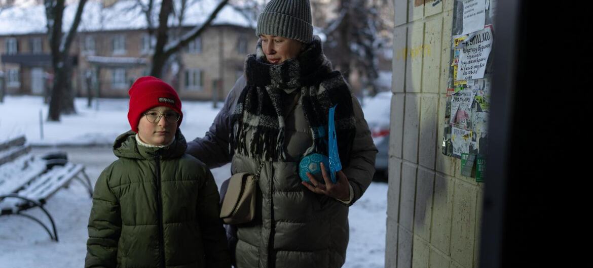 Женщина и ребенок, одетые в зимнюю одежду, стоят за зданием в снежной городской обстановке в Украине.
