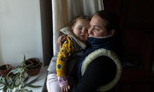 Una madre con una chaqueta de invierno sostiene a su niño dormido cerca de su pecho cerca de una ventana en un ambiente interior acogedor.