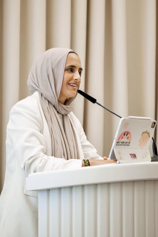 ماريام سوليكا، زعيمة الشباب في أهداف التنمية المستدامة التابعة للأمم المتحدة، تتحدث في مؤتمر بينما تجلس على المنصة مع لوحة.