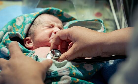 एक नवजात शिशु की नज़दीकी तस्वीर जो एक पैटर्न वाली कंबल में लपेटी हुई है, जिसे अस्पताल में एक स्वास्थ्य पेशेवर द्वारा स्टेथोस्कोप के साथ जांच की जा रही है।