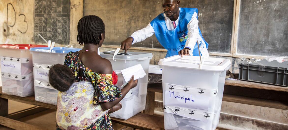 A woman carrying a child places a ballot into a transparent ballot box labeled 'Municipale' while an election official observes in a classroom setting.