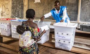 A woman carrying a child places a ballot into a transparent ballot box labeled 'Municipale' while an election official observes in a classroom setting.