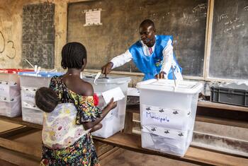 Une femme porteuse d'un enfant place un bulletin dans une boîte de vote transparente étiquetée "Municipale" tandis qu'un responsable électoral observe dans une salle de classe.