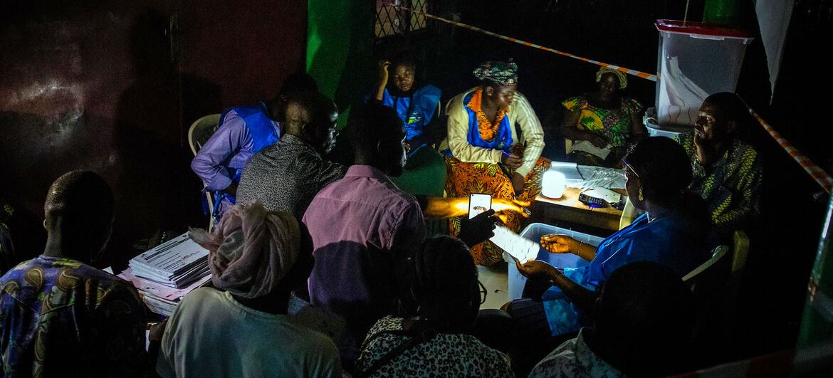 A group of people gathered around a table at night, counting ballots during an election under the light of a small lamp, with a ballot box in the background.