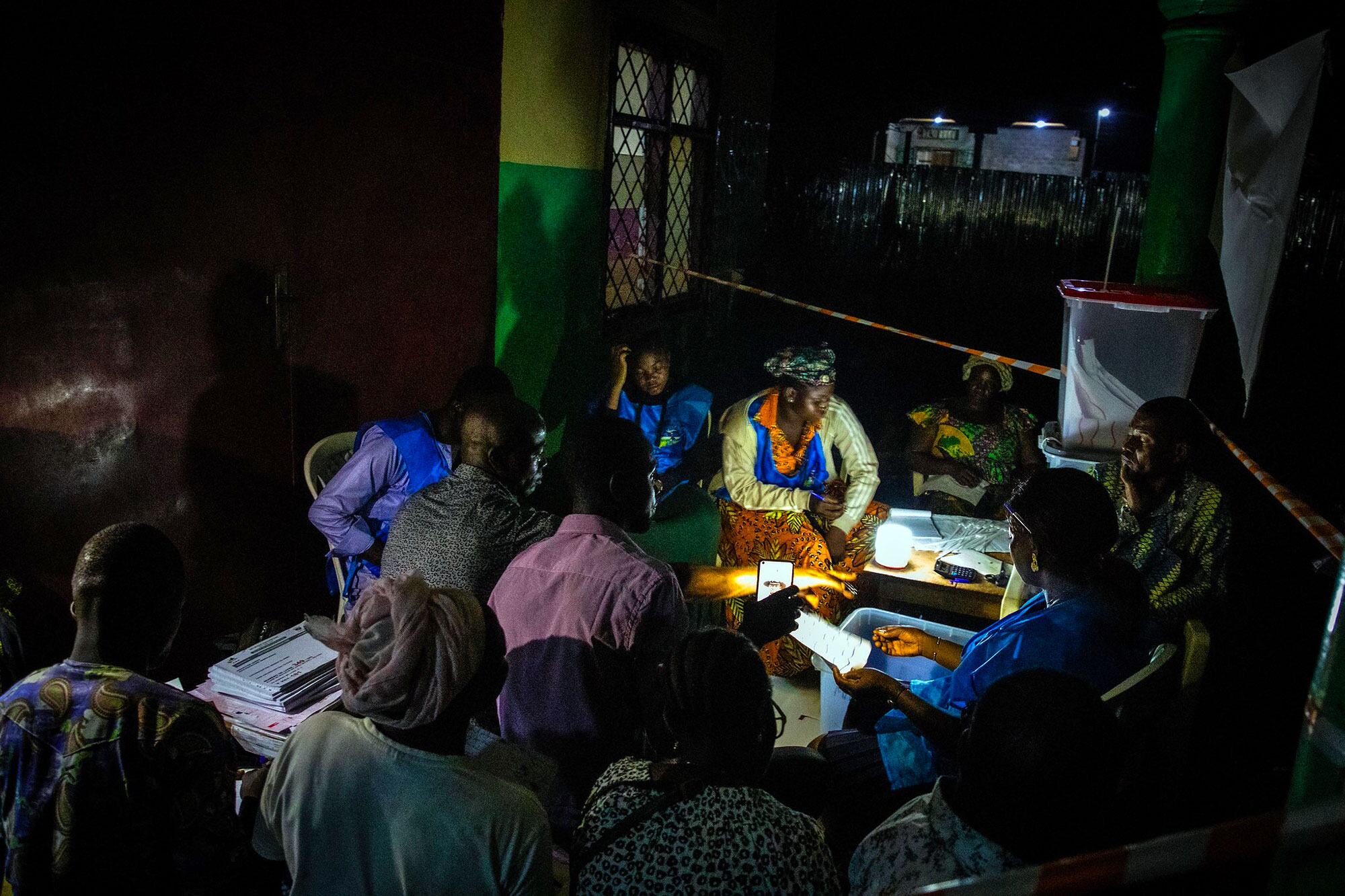 Un groupe de personnes s'est rassemblé autour d'une table la nuit, pour compter les bulletins lors d'une élection sous la lumière d'une petite lampe, avec une boîte à bulletins en arrière-plan.