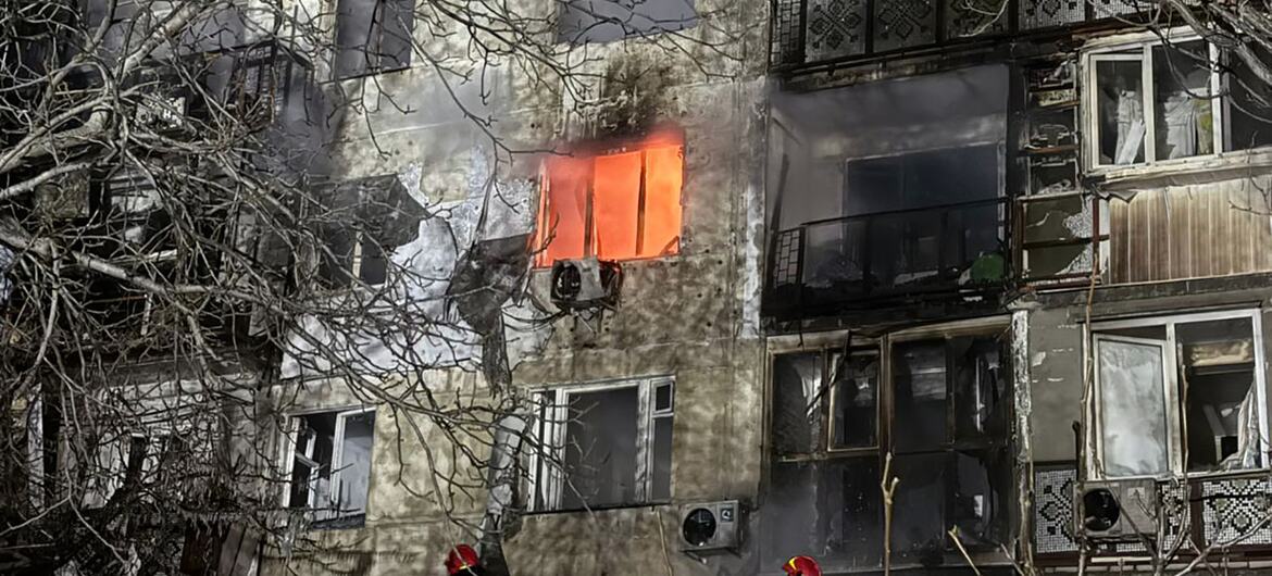 A damaged building in Odesa City with fire still burning in one of its windows, surrounded by bare trees. Rescuers and aid workers are present to assist affected civilians.