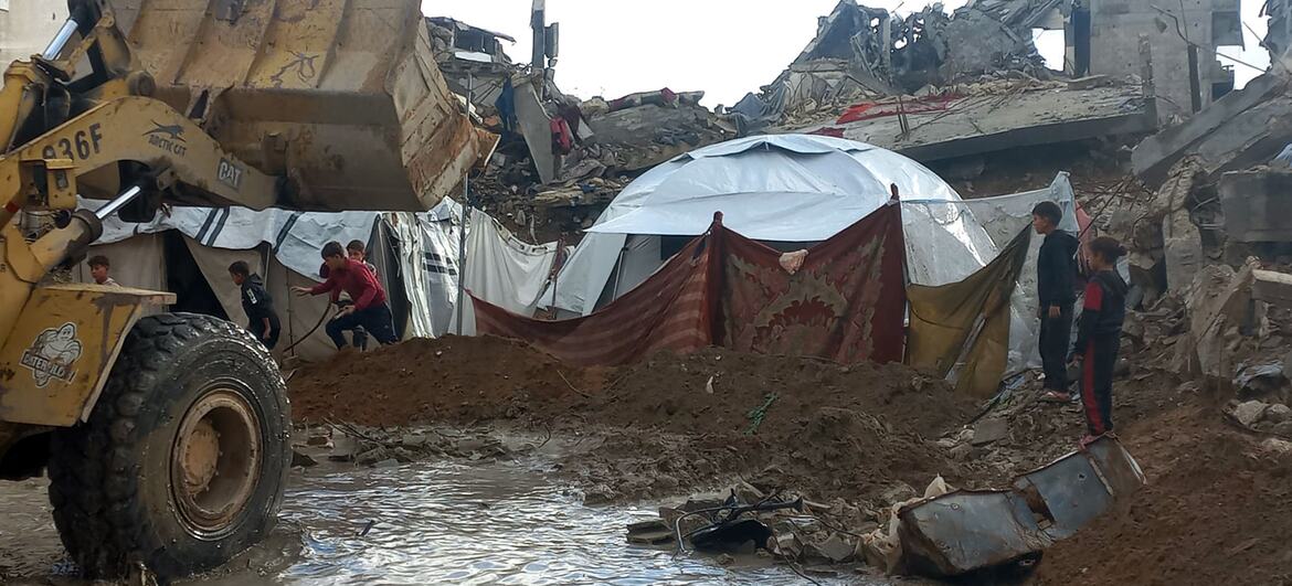 Los niños se encuentran cerca de un campamento de tiendas improvisadas en los escombros de edificios destruidos en la Franja de Gaza, ya que las fuertes lluvias e inundaciones han barrido la zona, con un vehículo amarillo de construcción parcialmente sumergido en el agua fangosa.