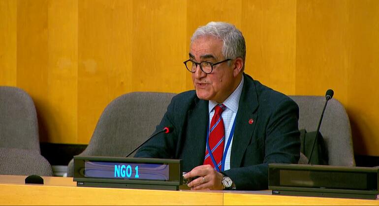 رجل يحمل نظارات وملابس يتحدث في اجتماع للأمم المتحدة، يجلس على مكتب مع لوحة "منظمة غير حكومية 1".