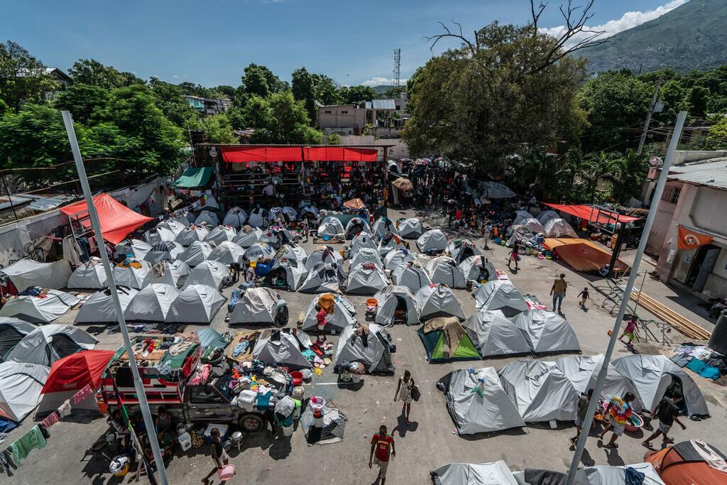 Uma vista aérea de um campo de refugiados lotado no Haiti, com numerosas tendas cinzas instaladas em uma área urbana. Pessoas são vistas caminhando ao redor e algumas são reunidas sob um dossel vermelho. Montanhas e árvores são visíveis no fundo.