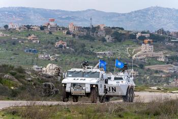 وركبتان مدرعتان تابعتان لليونيفيل تحملان أعلام الأمم المتحدة وأفرادا مسلحين يقومون بدورية من خلال مشهد ريفي مائي تلي به مبان مبعثرة في الخلفية.