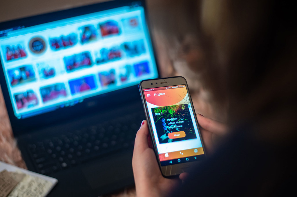 Uma pessoa segura um smartphone exibindo a tela de um programa, com um laptop mostrando uma galeria de imagens ao fundo.