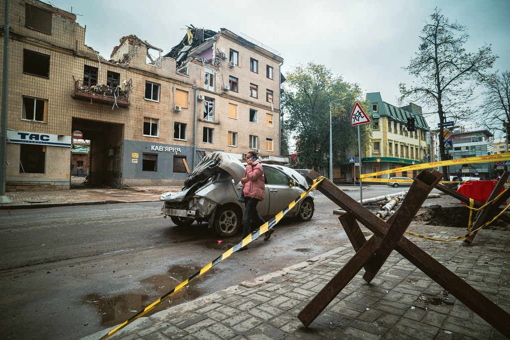 Un automóvil dañado y una mujer de pie en una calle del centro de Járkov, Ucrania, con un edificio destruido al fondo.