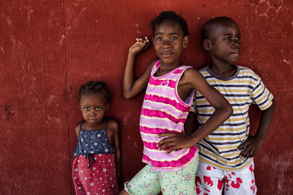Watoto watatu, wasichana wawili na mvulana mmoja, wakipiga picha dhidi ya ukuta mwekundu katika kambi ya watu waliokimbia makazi yao nchini Haiti.